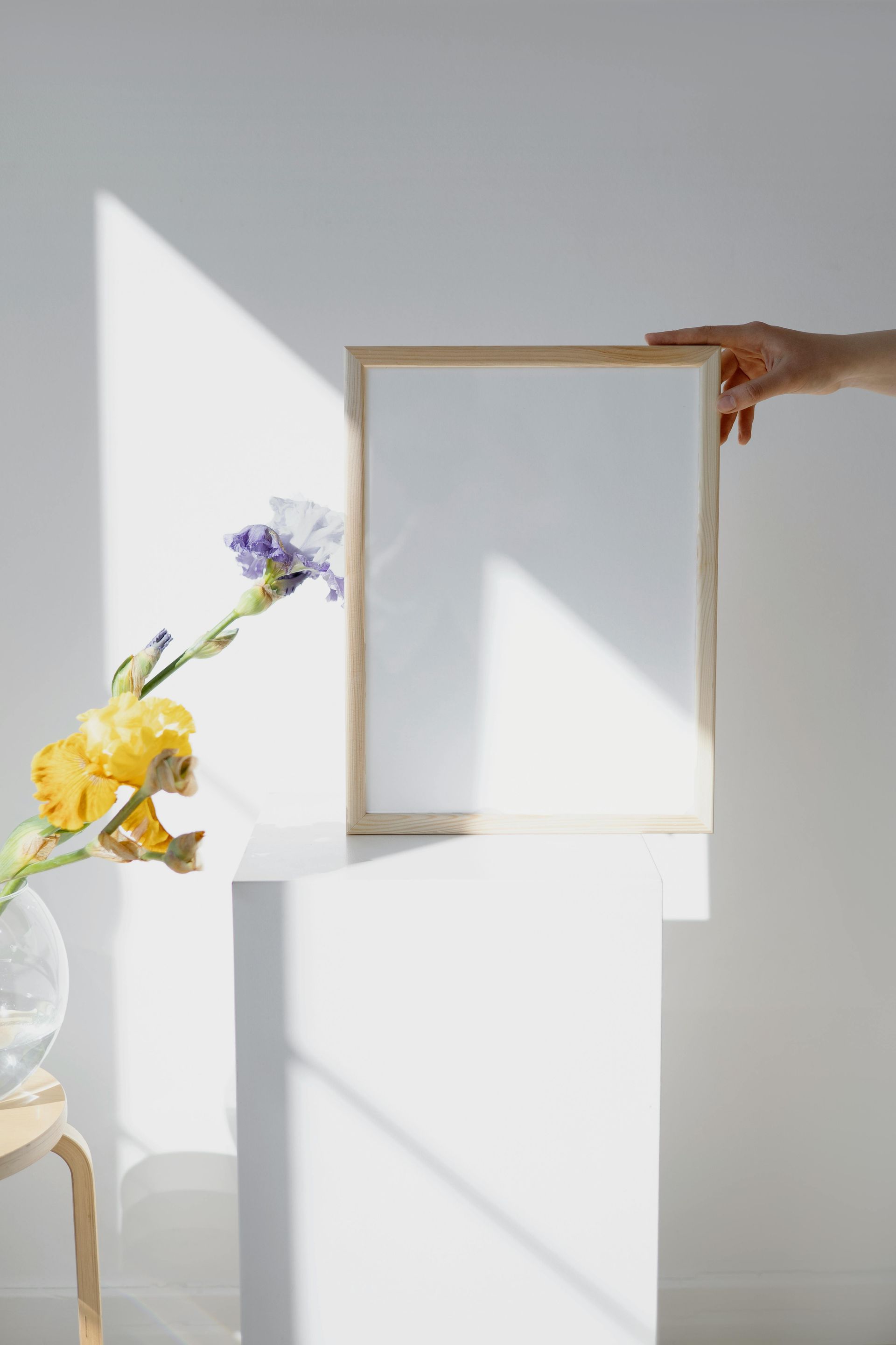 Hand holding a blank frame on a white pedestal, flower arrangement to the left, sunlight casting shadows.