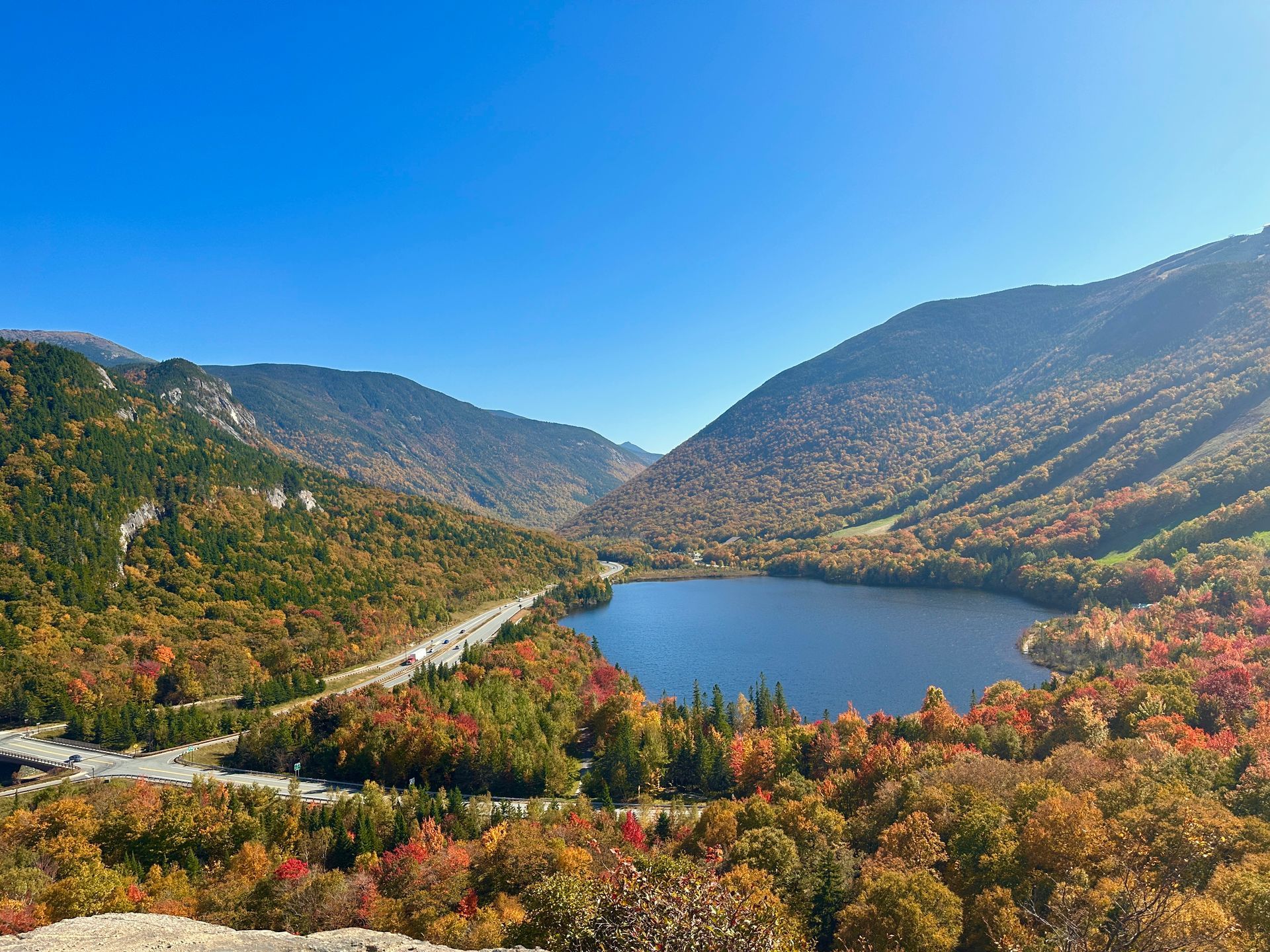 fall view in franconia notch state park featuring mountains, a lake, a blue sky, and a road with cars, taken on a hot day in october, taken from a viewpoint at artists bluff hiking trail