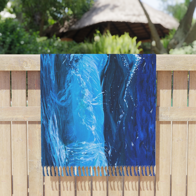 boho beach blanket with blue painting of waves on it and tasseled edges draped over a light wooden fence outside with trees in the background