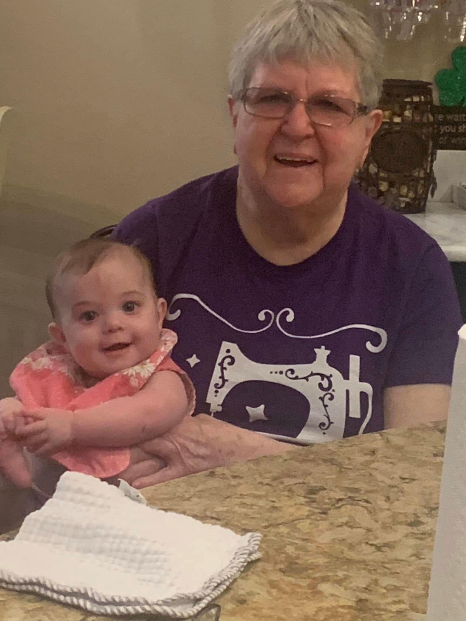 Woman in purple shirt holds baby, both smiling at the camera. They sit at a table.