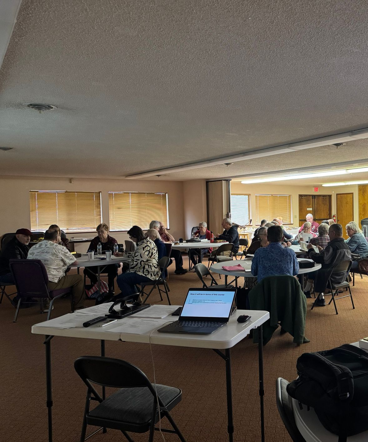 Group of people seated at tables in a brightly lit room, looking at laptops and papers, possibly a meeting or workshop.