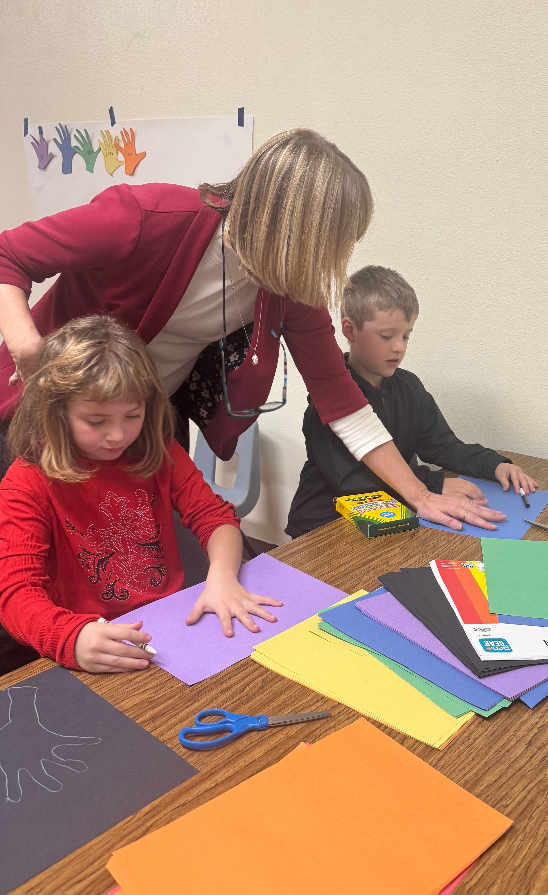 Teacher assisting two children with craft project using colored paper and scissors.