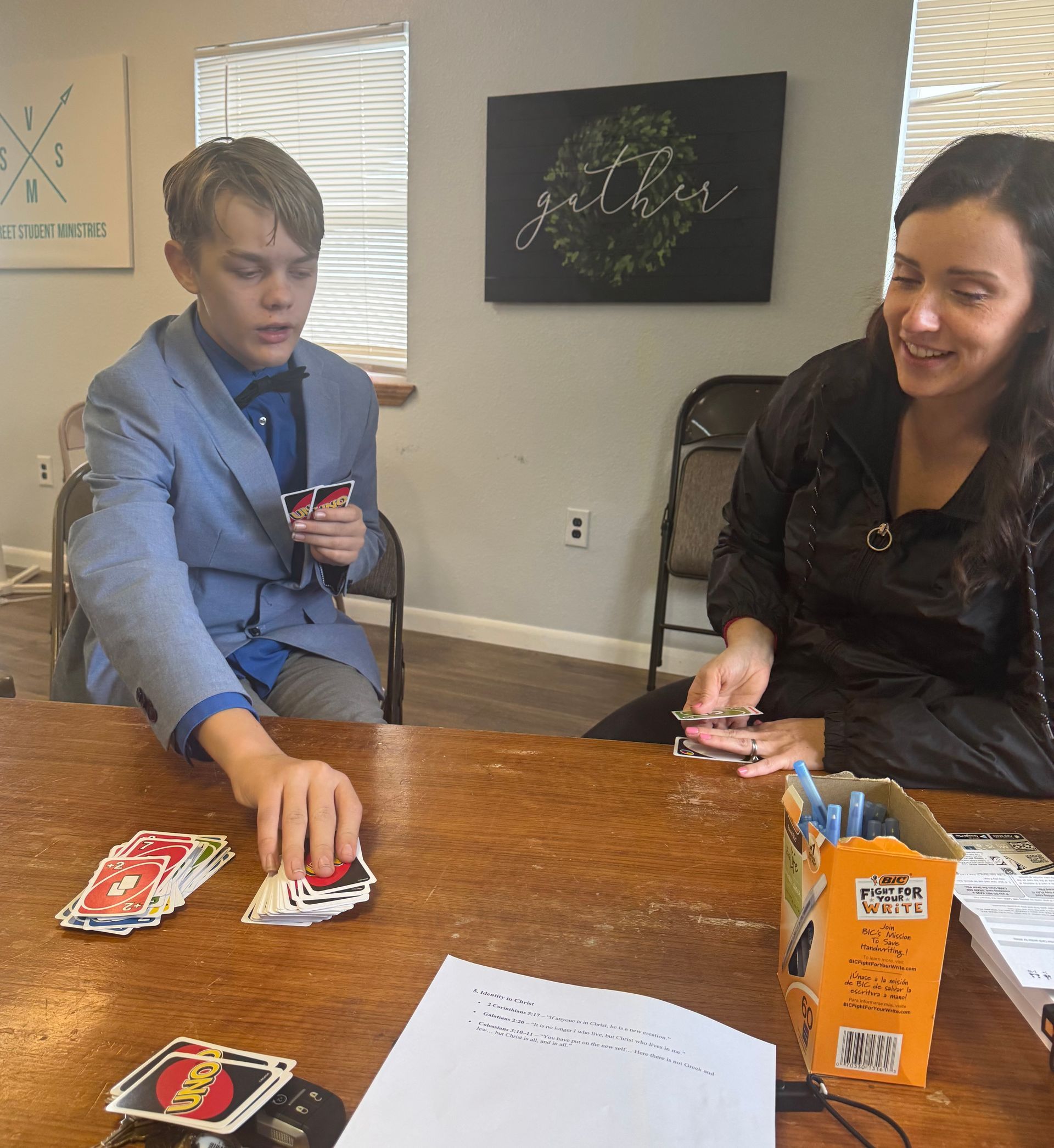 Person in blue suit plays cards with woman at a wooden table. A game is in progress indoors.