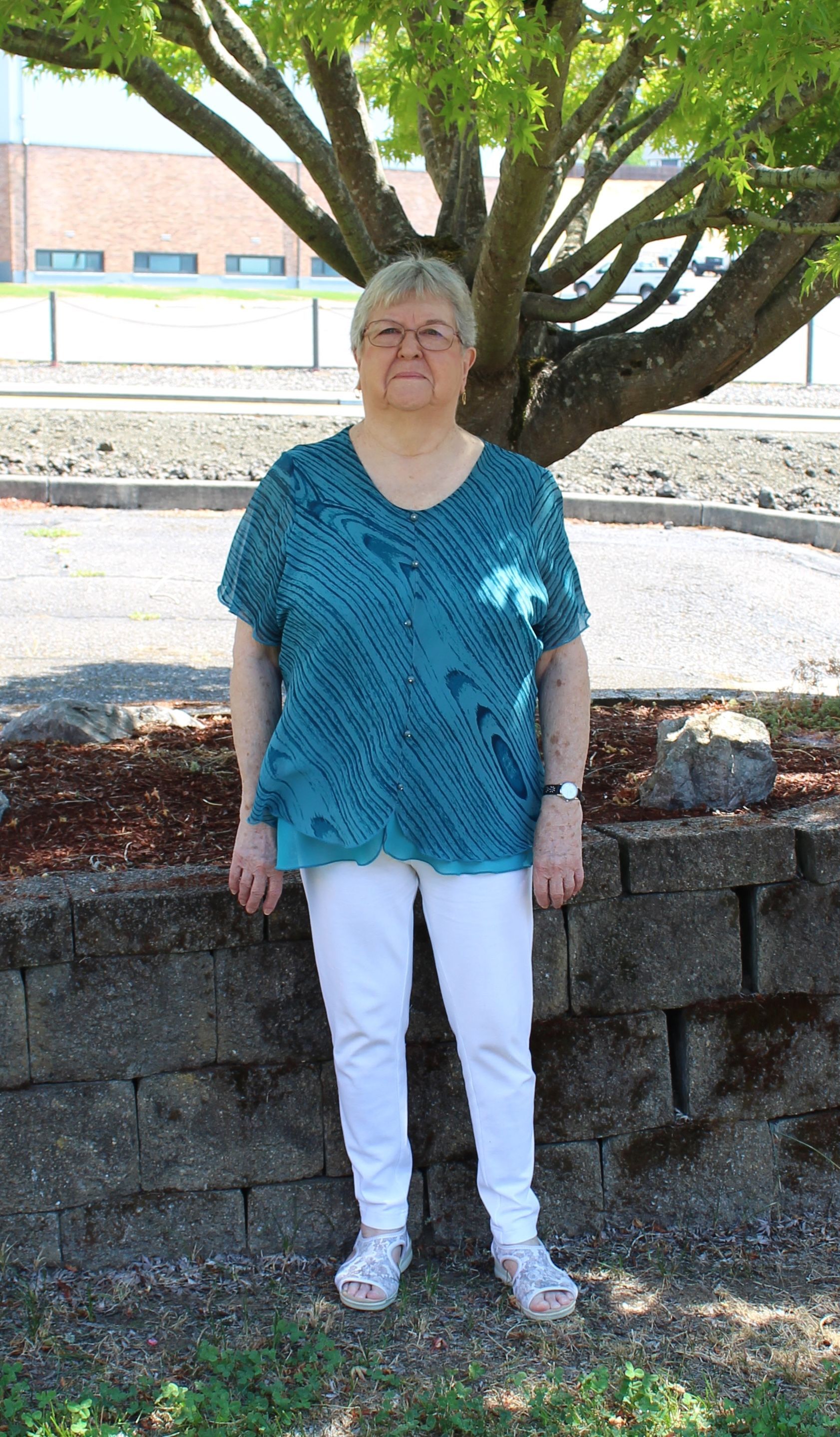 Woman in blue top and white pants standing near a tree and a stone wall.