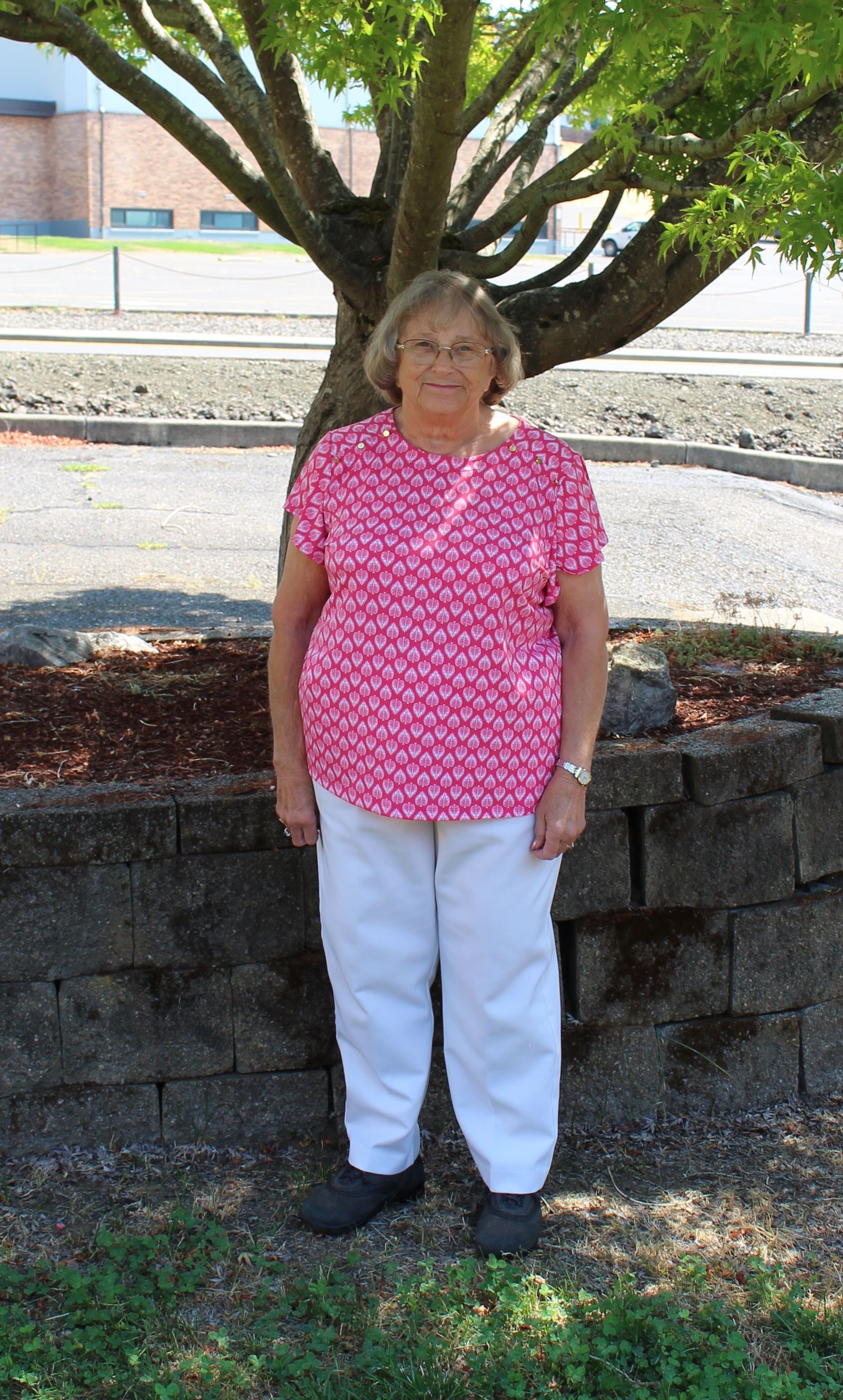 Woman in pink patterned shirt and white pants stands under a tree in front of a building.