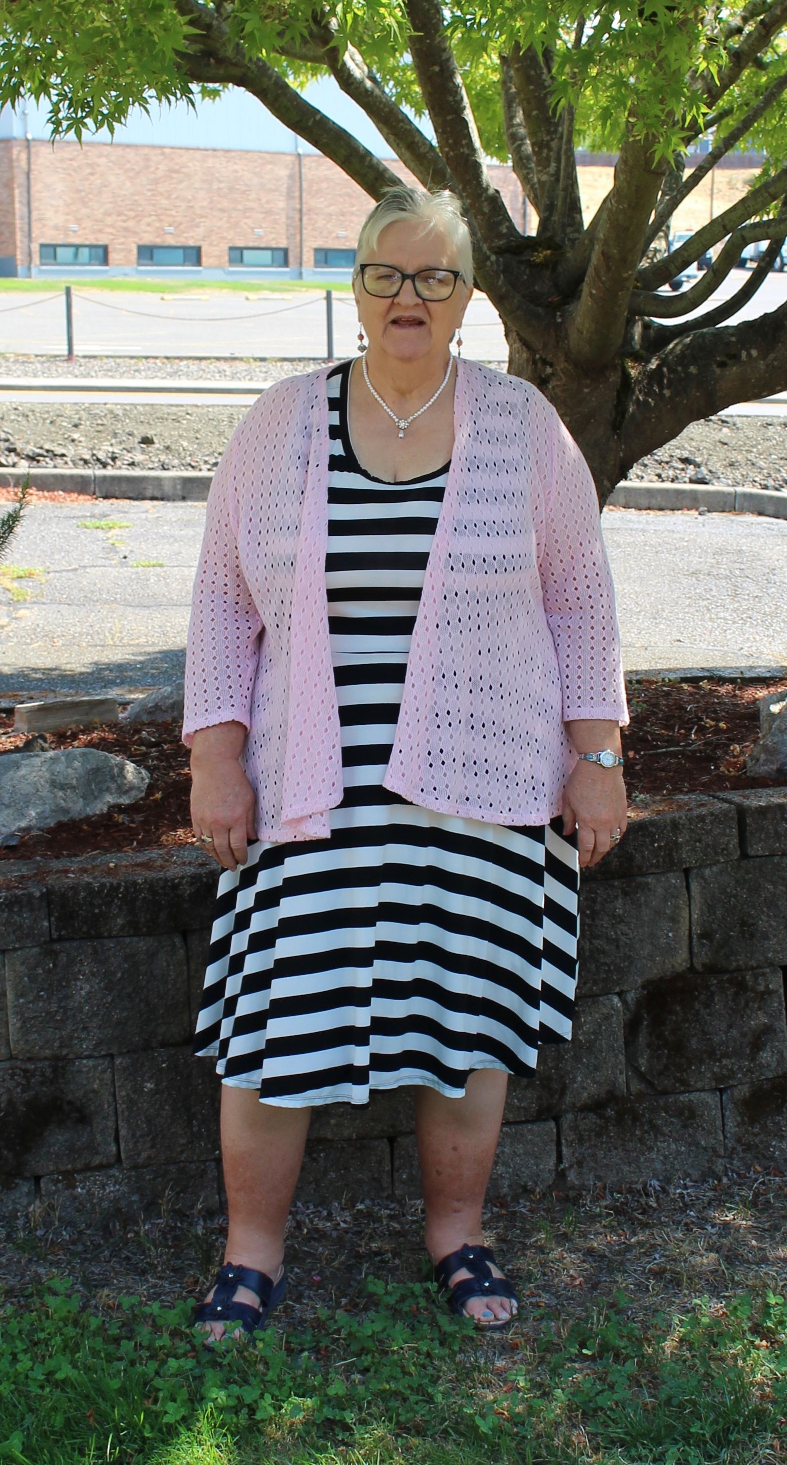 Woman in striped dress and pink cardigan stands near a tree in front of a building.