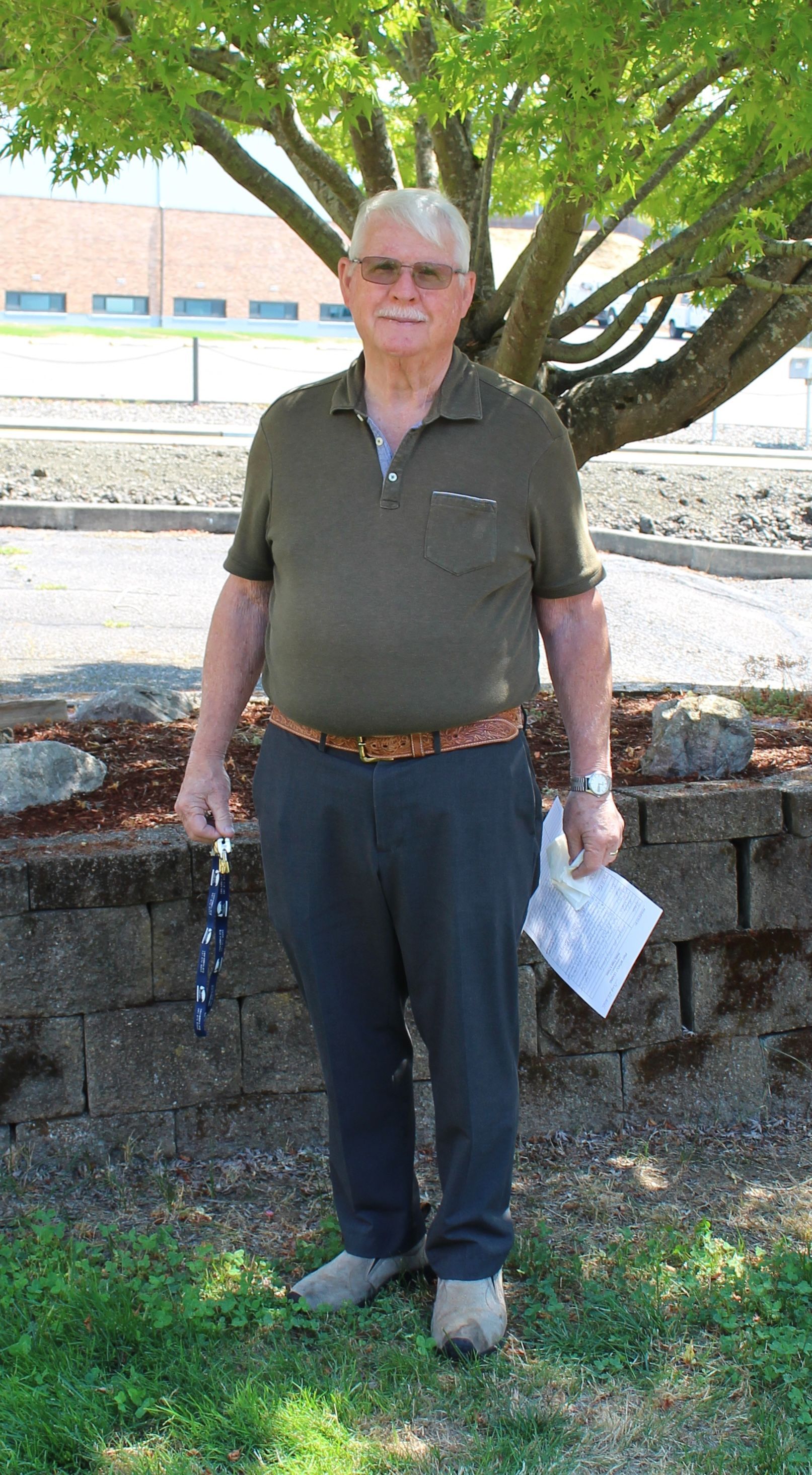Man standing near a tree, wearing a green shirt and grey pants, holding keys and a paper.