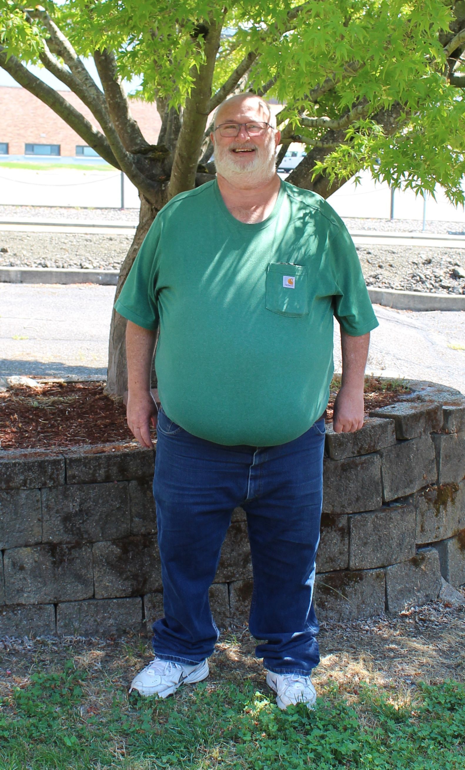 Man in green shirt and blue jeans standing outdoors, smiling, near a tree and stone wall.