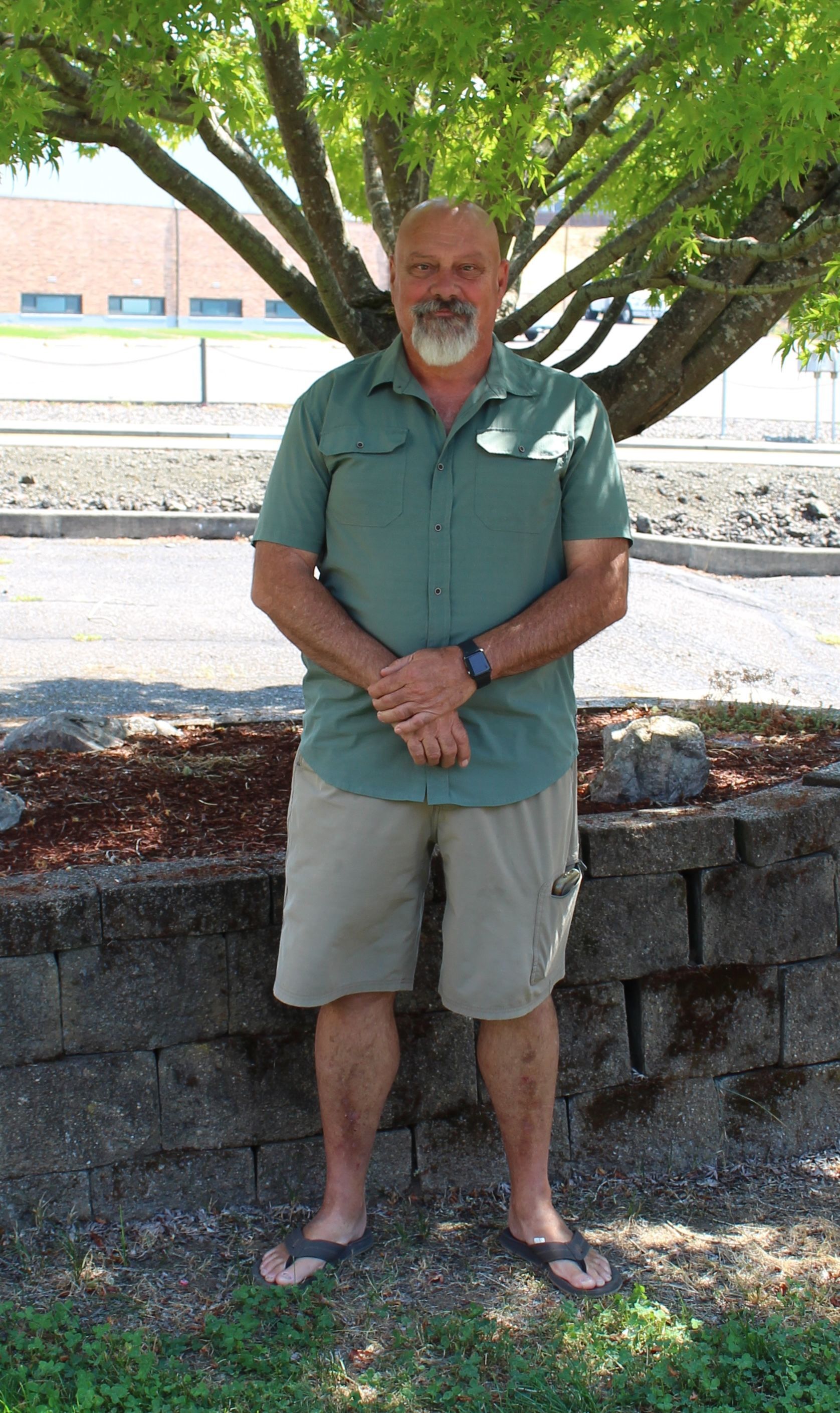 Man in green shirt and khaki shorts stands near a tree, hands crossed. Outdoors, sunny day.