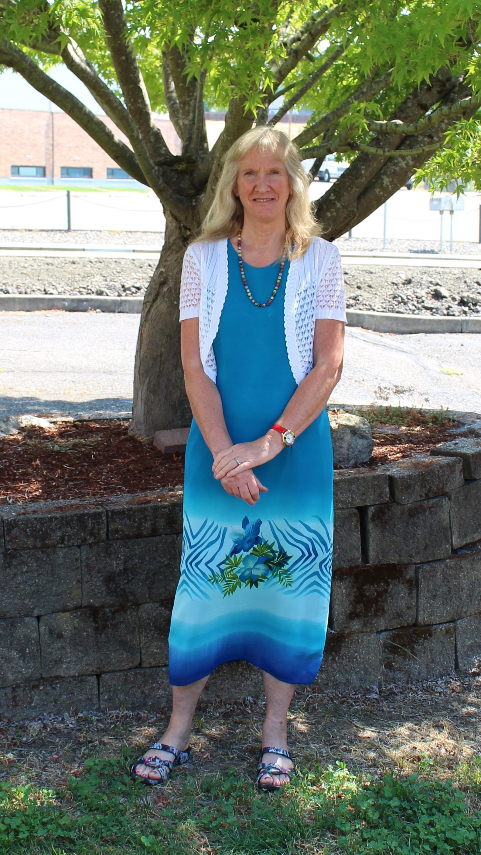 Woman standing in front of a tree, wearing a blue dress and white sweater.