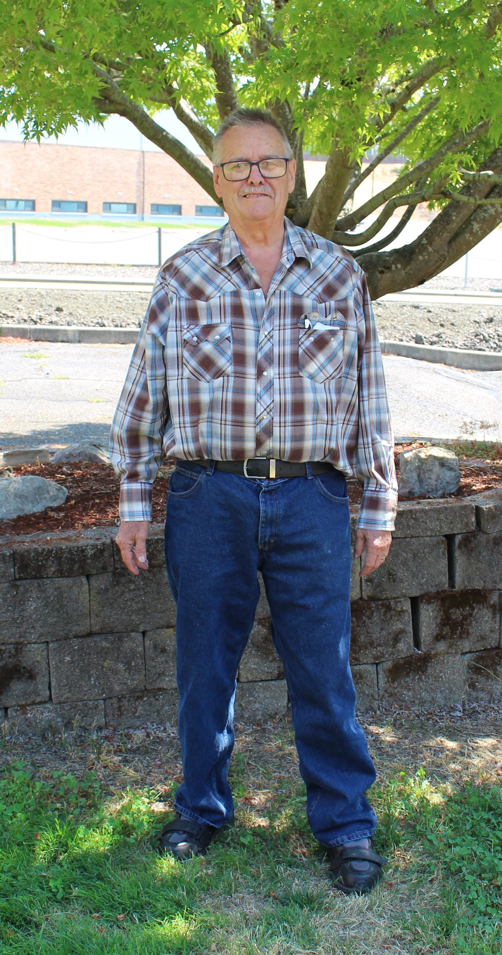 Man in plaid shirt and jeans stands outdoors, near a tree and brick wall.