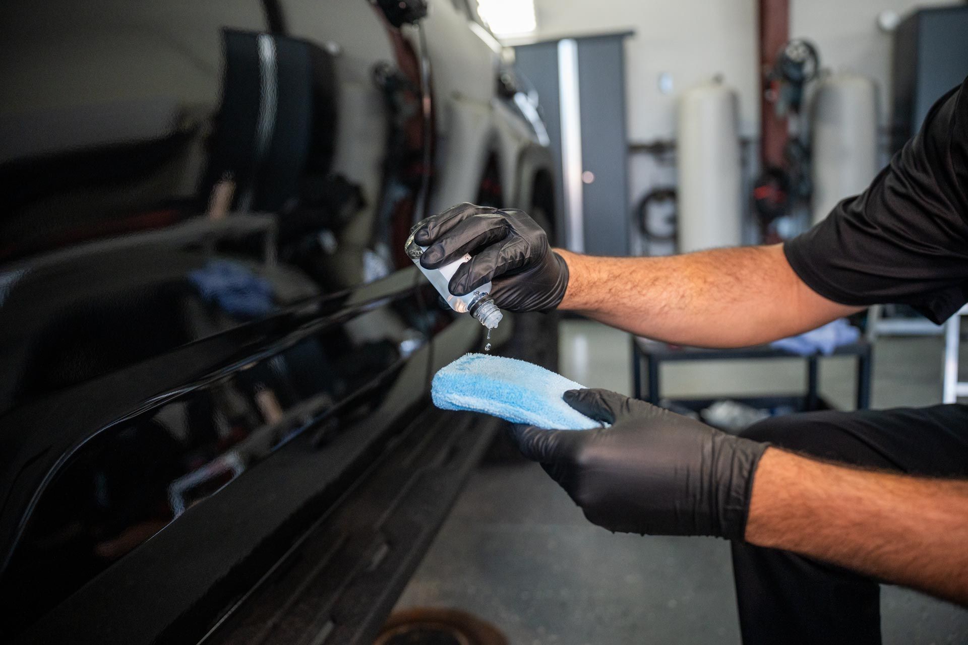 Person applying a liquid product to a car's black paint with a blue applicator sponge, indoors.