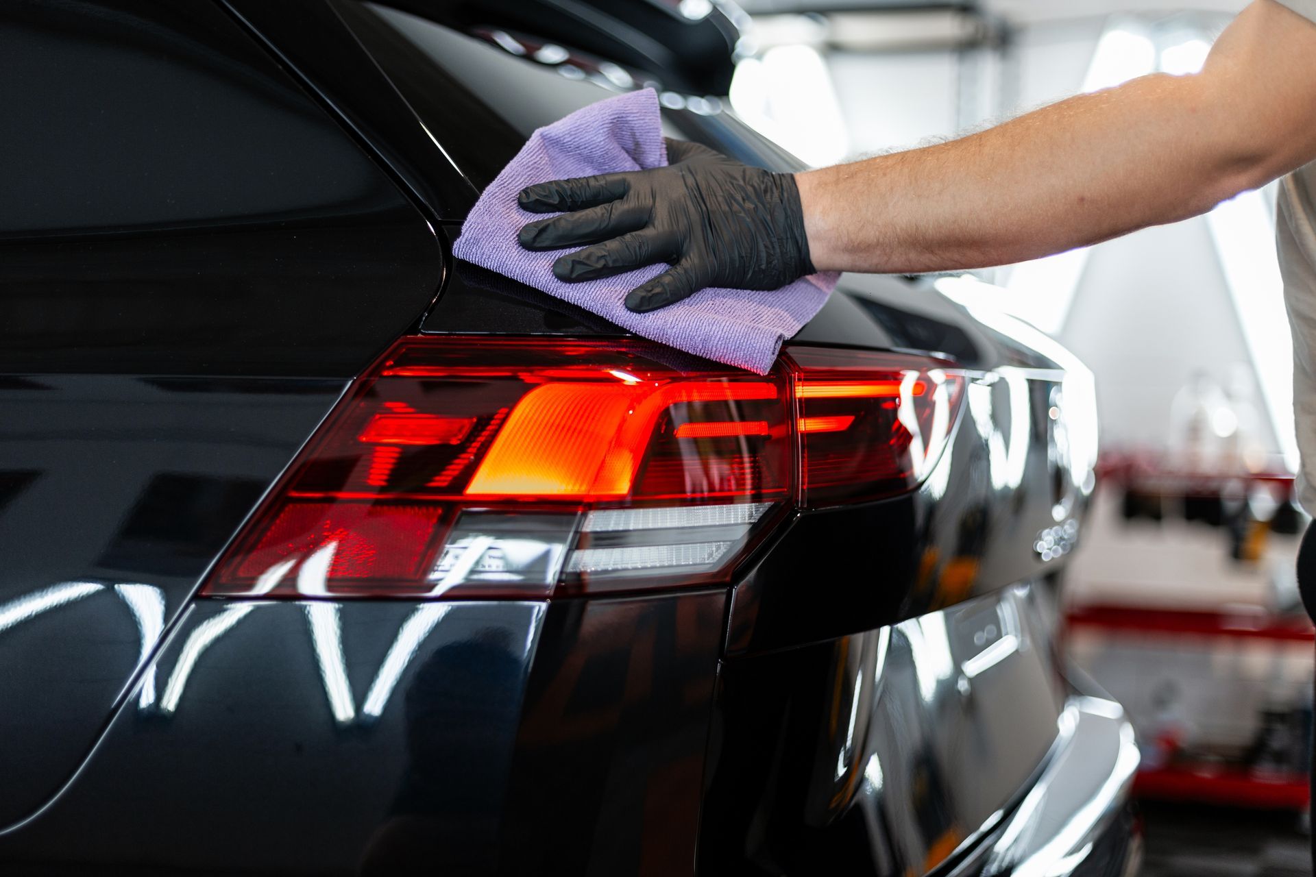 Person wearing a black glove wiping a black car's taillight with a purple cloth in a garage.