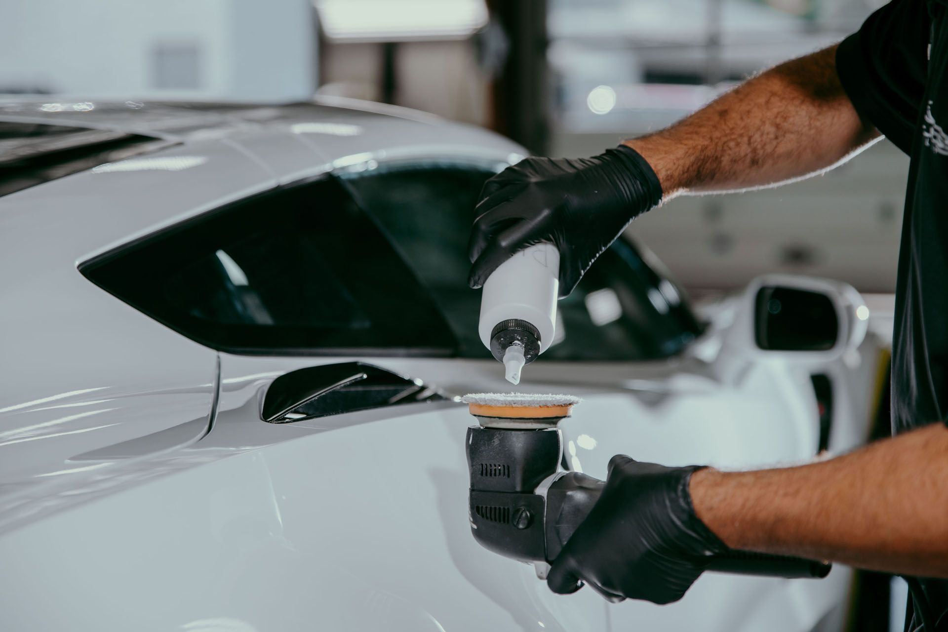 Person in black gloves polishing a white car with a power tool, indoors.