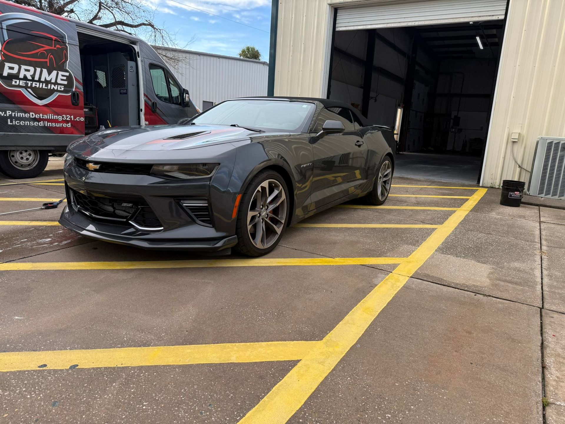 Dark gray Chevy Camaro parked in front of a garage with a service van nearby.