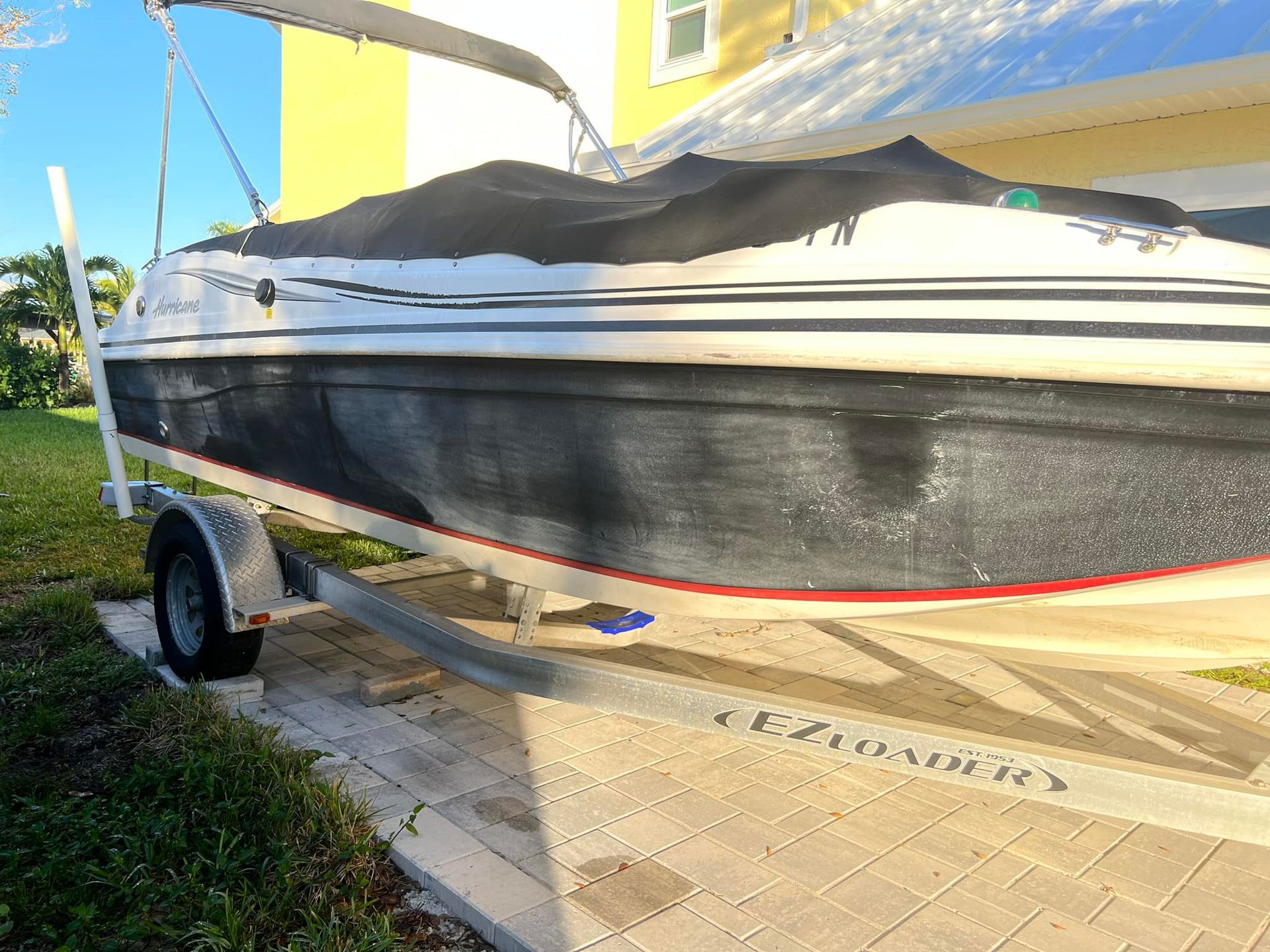 Boat on trailer, mostly black hull with white and red accents, under a black cover, parked on a concrete path.