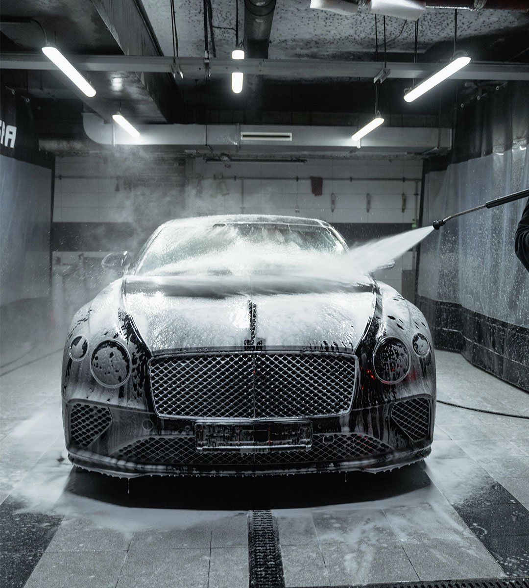 Bentley being washed at a car wash, covered in soapy foam, under bright lights.