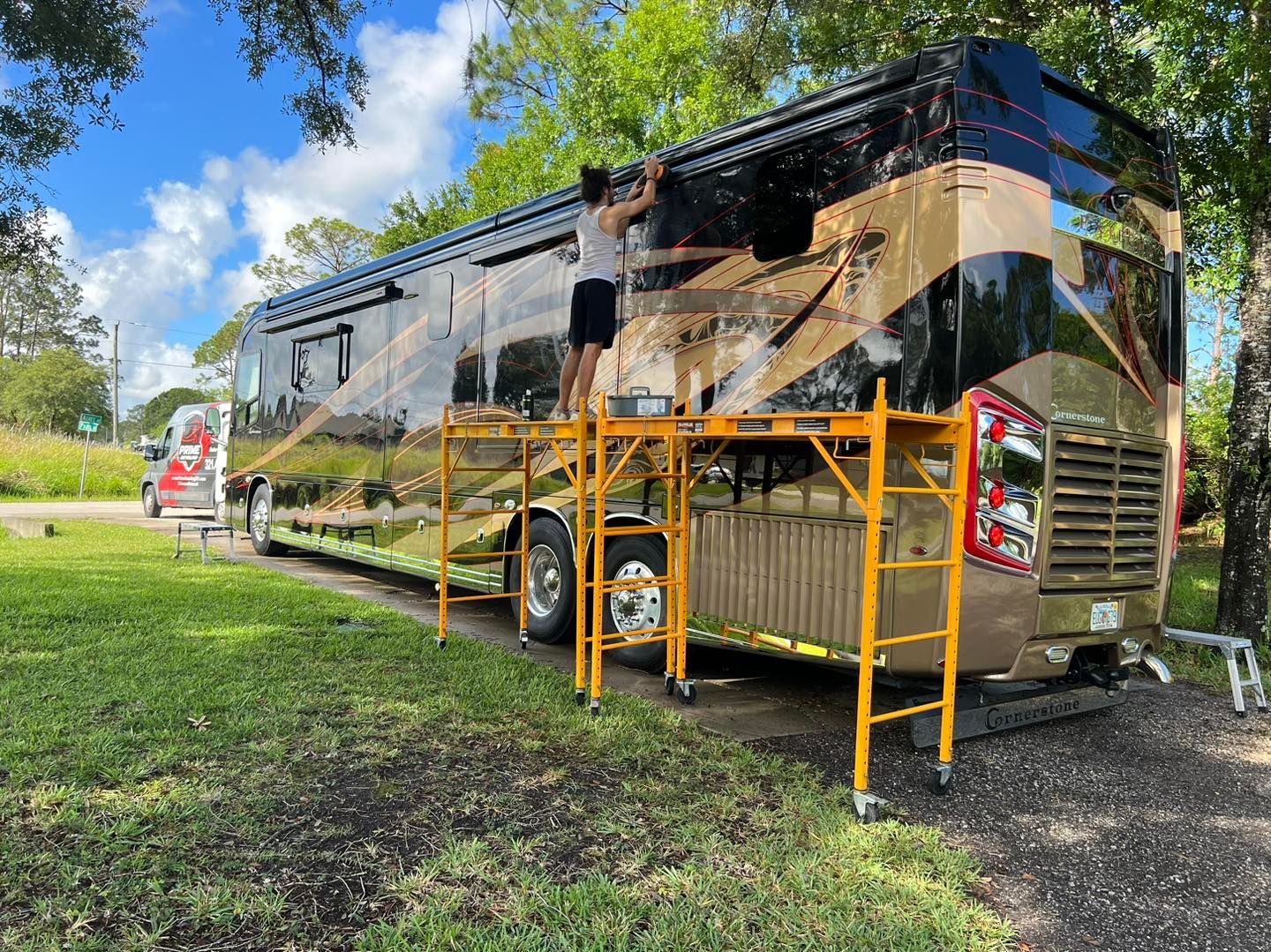 Person on scaffolding working on a large RV with gold and black paint, parked on grass.