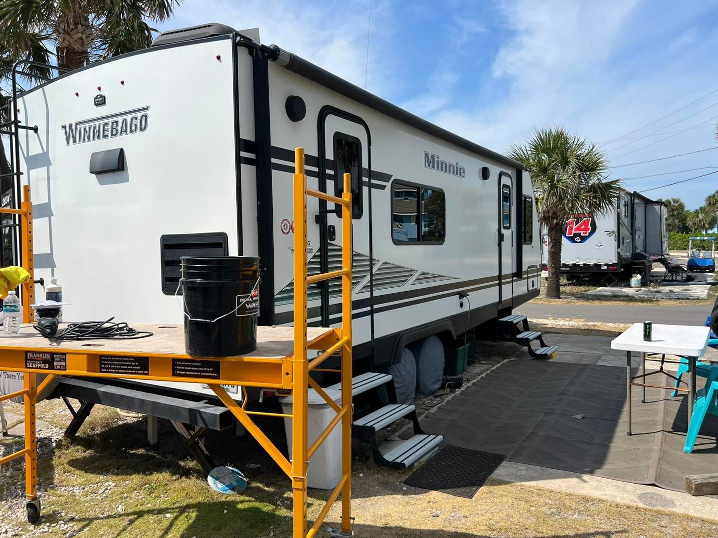 A white Winnebago travel trailer is parked outside with scaffolding, a bucket, and a table.