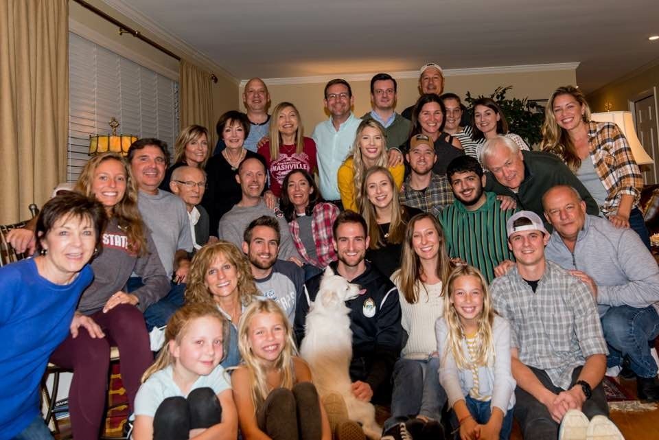 Six smiling people pose on a porch, leaning together in a close family-style group selfie.