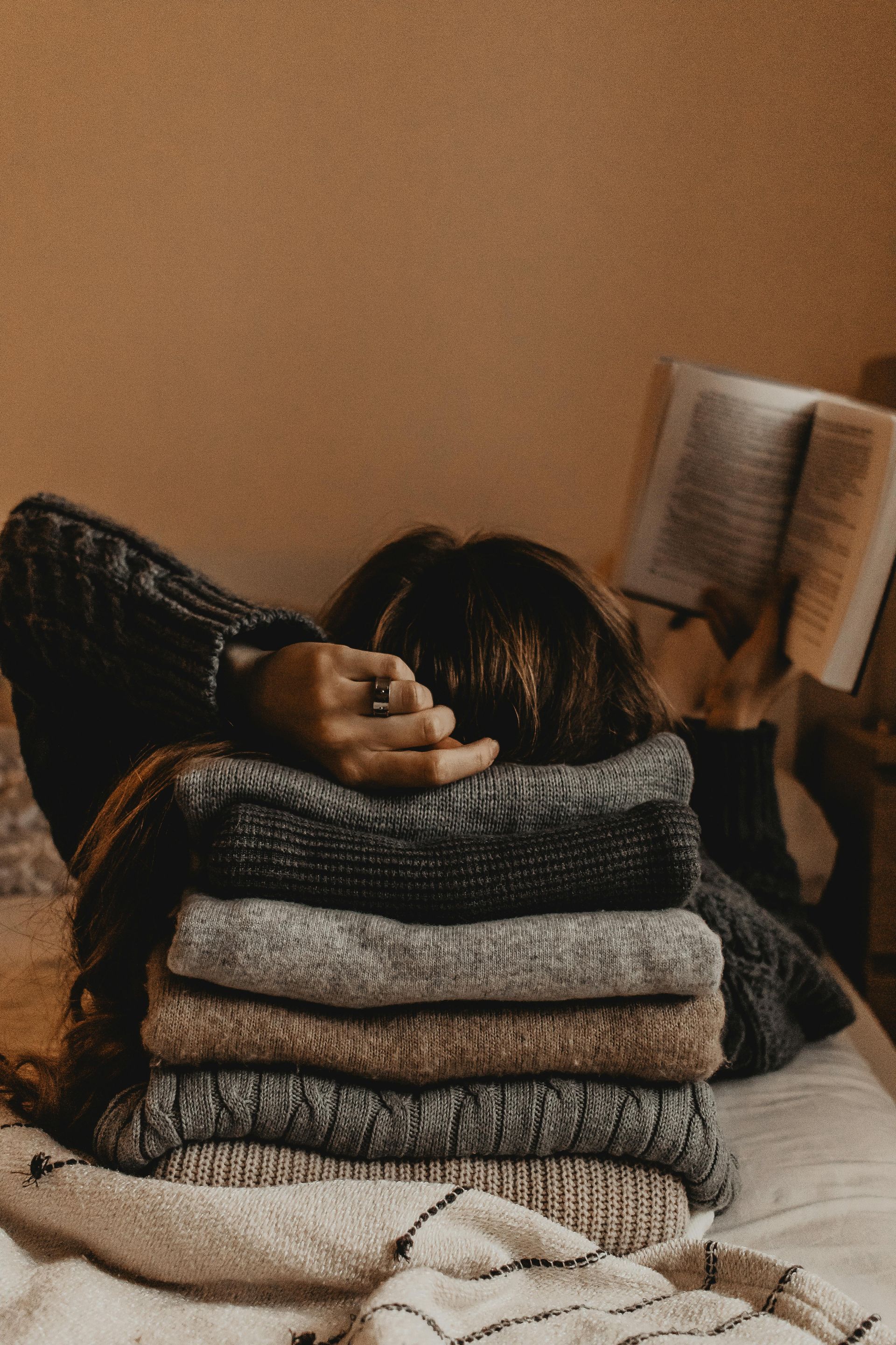 A woman is laying on a bed with a stack of sweaters and a book.