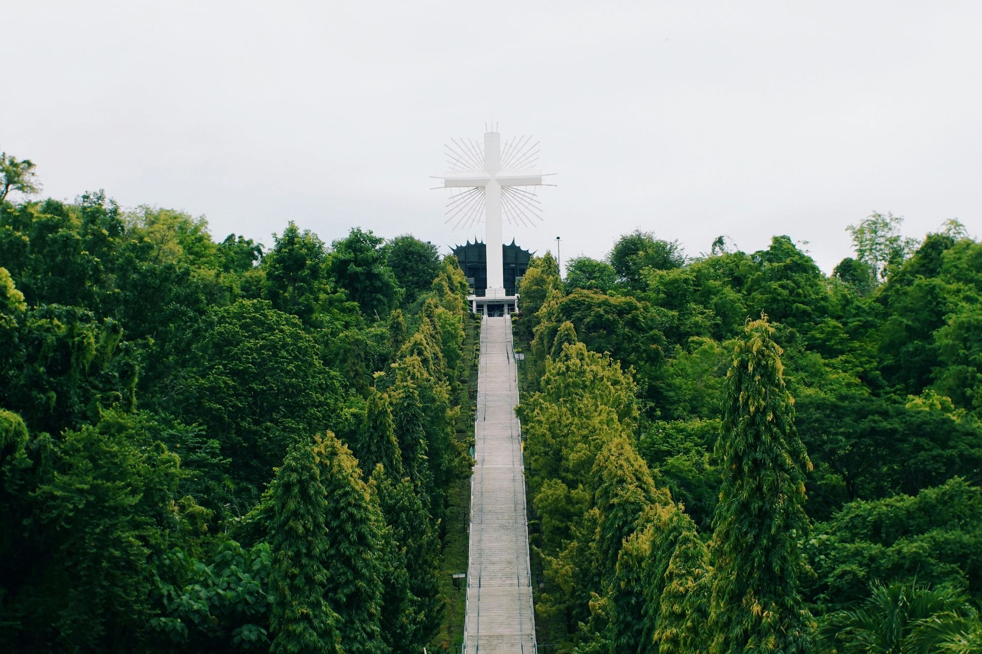 A long paved path leads to a large white cross amidst dense green trees under a cloudy sky.