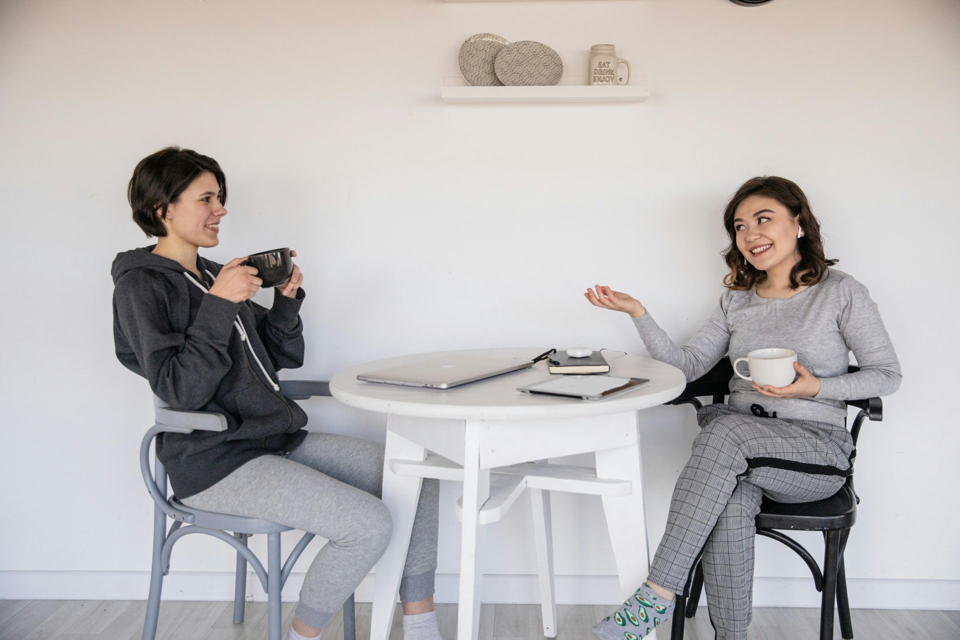 Two women in casual clothing sitting at a white table, talking. One drinks coffee, the other gestures. White room.