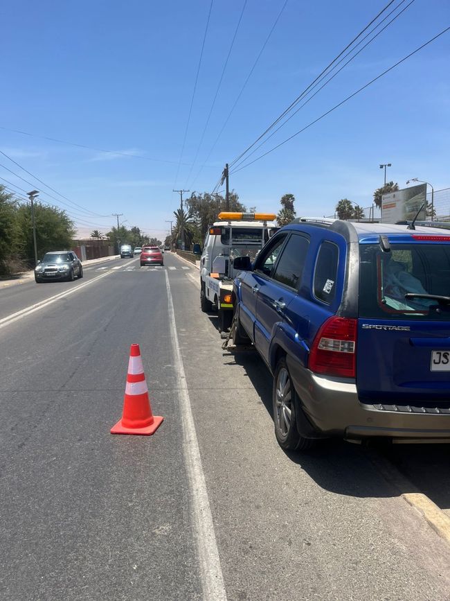 Una grúa está estacionada en el arcén de una carretera de dos carriles, remolcando un SUV azul, con un cono de tráfico naranja colocado cerca.