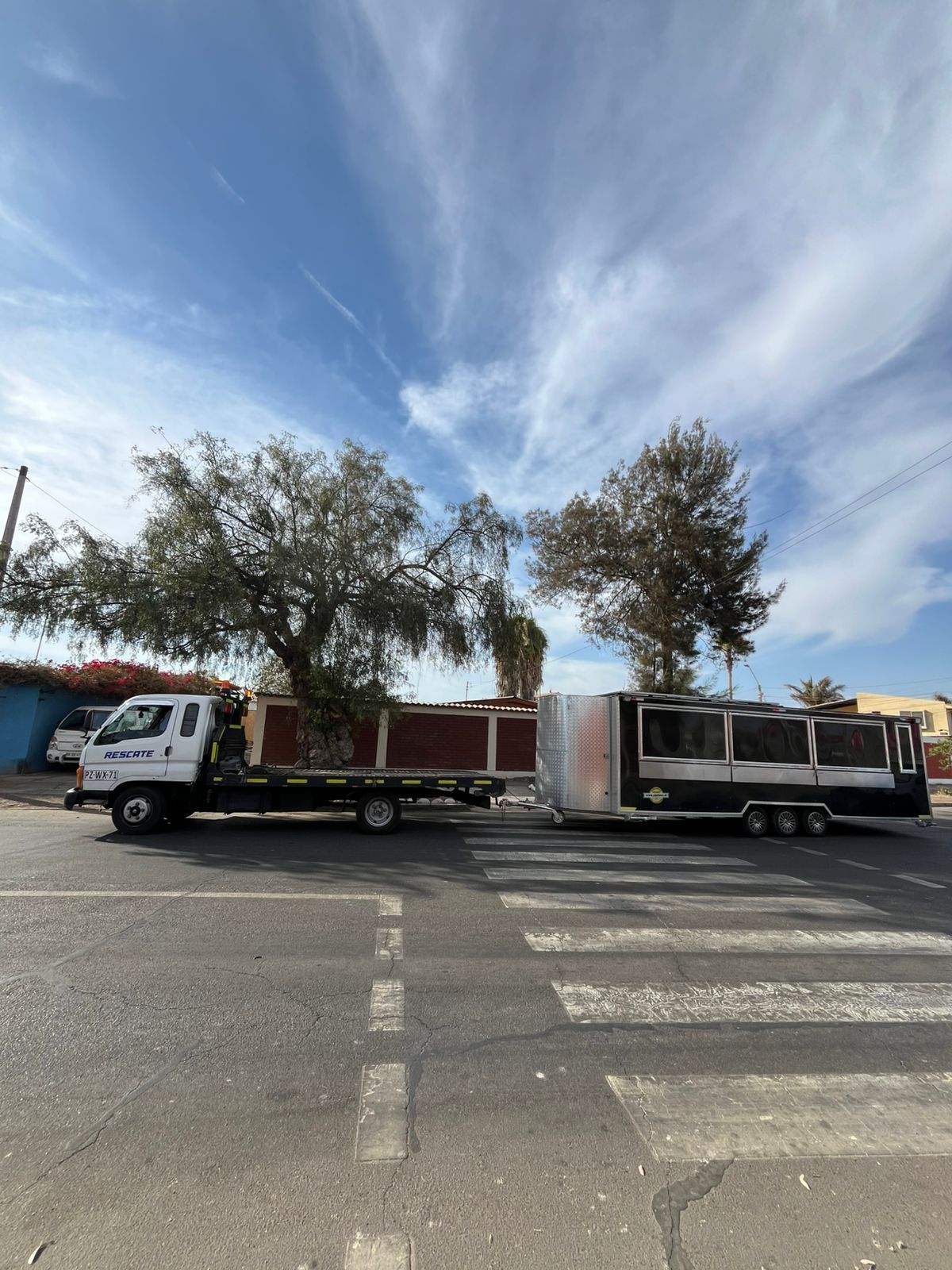 Una grúa de plataforma blanca arrastra un remolque largo, negro y plateado, a través de un paso de peatones en una carretera asfaltada bajo un cielo azul.