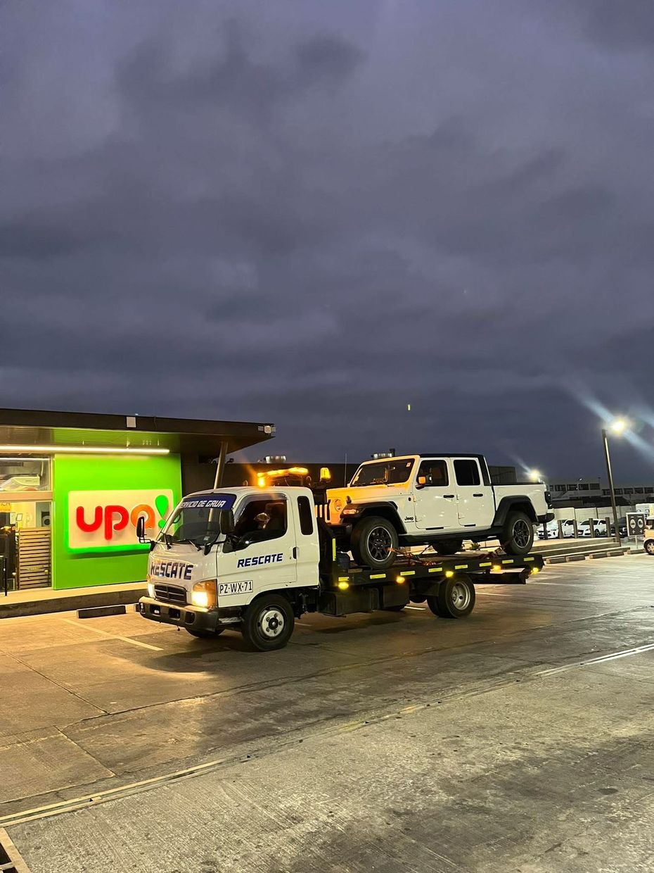 Una grúa de plataforma transporta una camioneta blanca estacionada frente a un edificio verde de la tienda UPN al anochecer.