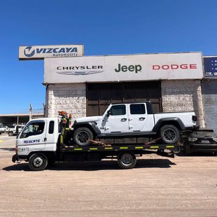 Una camioneta Jeep Gladiator blanca se encuentra asegurada sobre una grúa de plataforma frente a un concesionario de Chrysler, Jeep y Dodge.