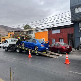 Un Subaru WRX azul está siendo subido a una grúa blanca en una calle de la ciudad, señalizada con dos conos de tráfico naranjas.