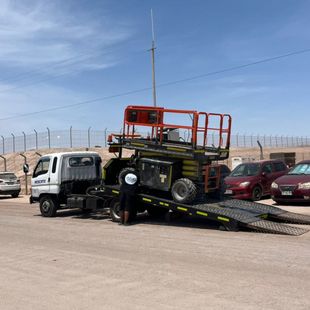 Un camión blanco de plataforma está estacionado en un aparcamiento, cargado con una plataforma elevadora industrial de tijera de color naranja, bajo un cielo azul claro y brillante.