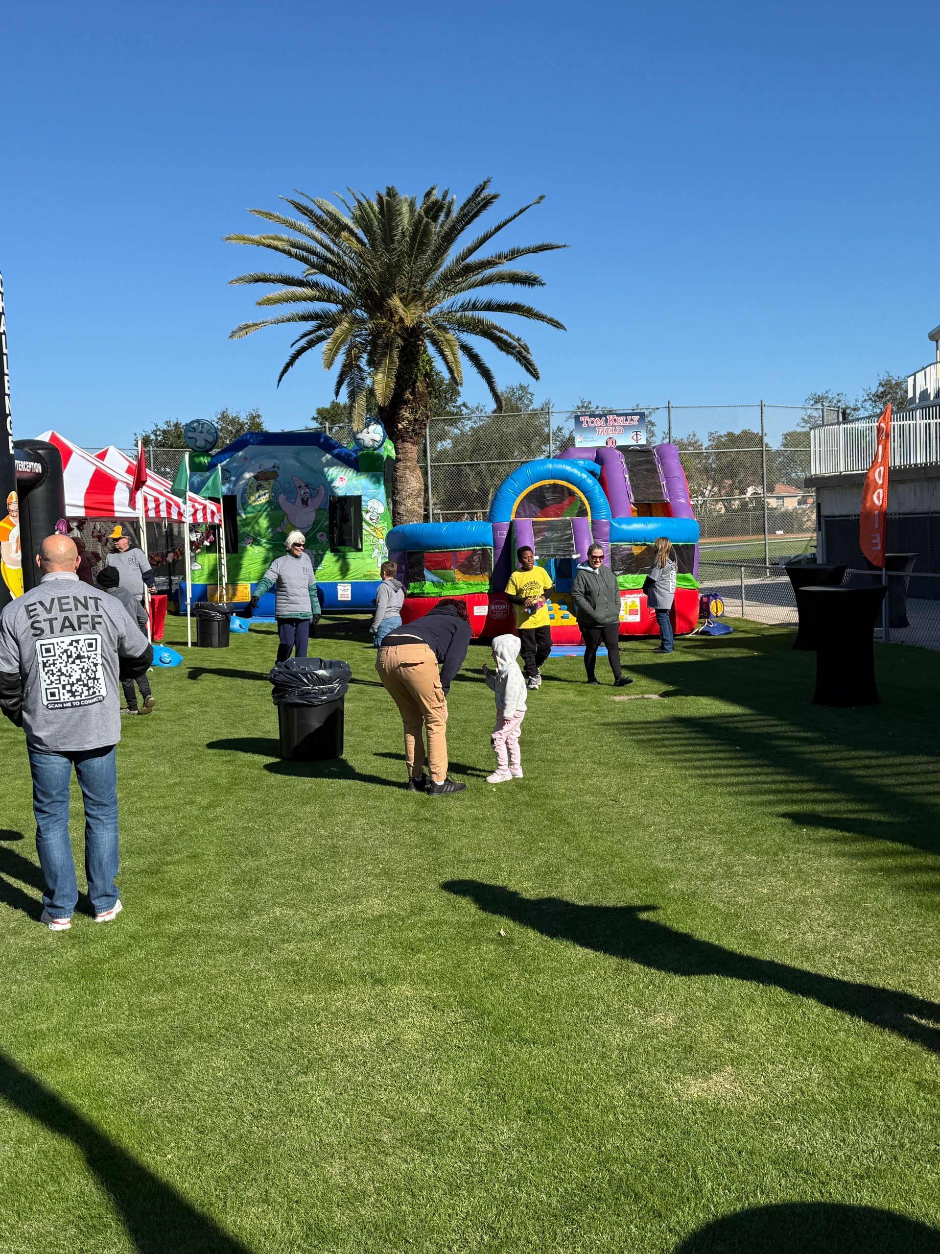 People at an outdoor event with bounce houses, a palm tree, and green grass on a sunny day.