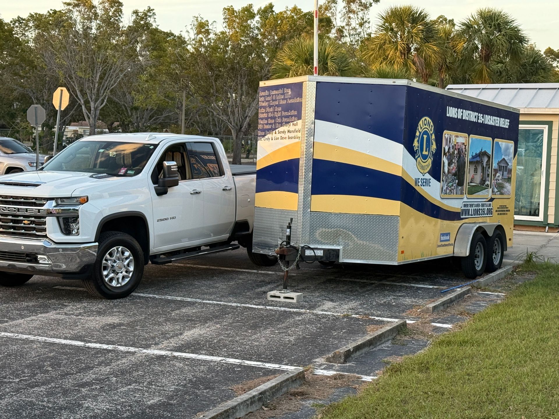 White truck towing a trailer with blue and yellow Lions Club graphics in a parking lot.