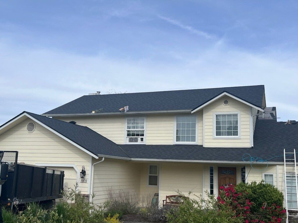 A large white house with a black roof and a dumpster in front of it.