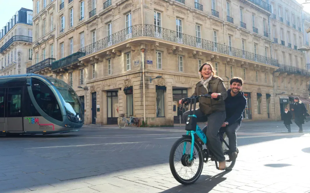 Eine junge Frau  fährt ein Leihfahrrad in Paris, auf dem Gepäckträger fährt ein junger Mann mit, lachend