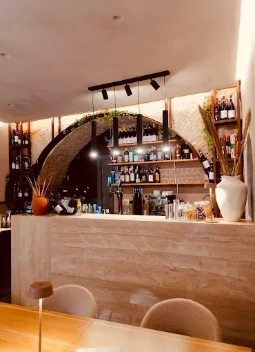 Interno del bar con scaffali ad arco, bottiglie, bevande e illuminazione, con una tavolozza di colori neutri.