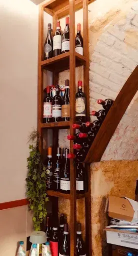 Scaffale di legno pieno di bottiglie di vino in un ristorante, contro un muro di mattoni beige e una pianta verde.