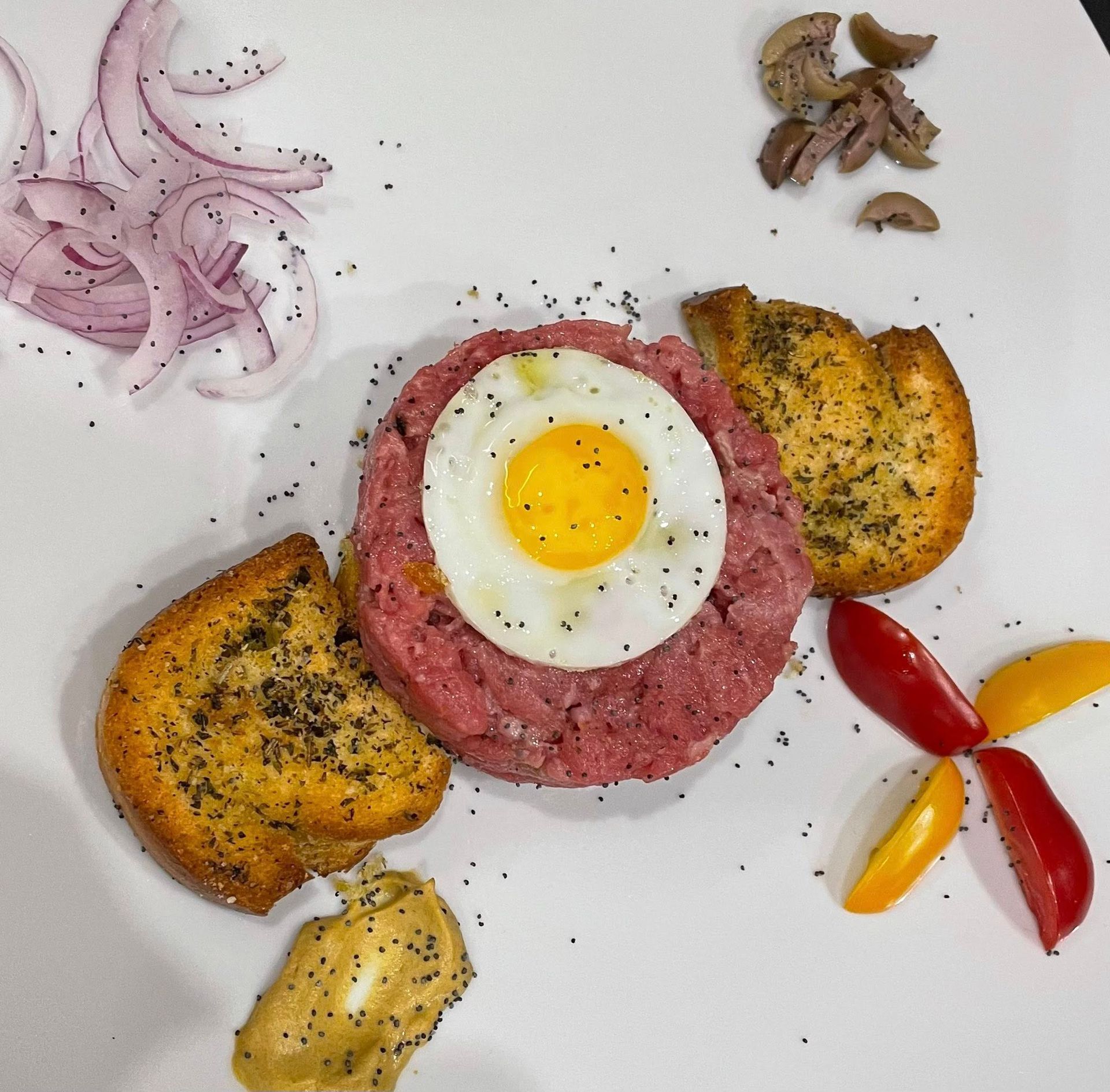 Tartare di manzo con uovo fritto, crostini, cipolla, olive e senape su un piatto bianco.