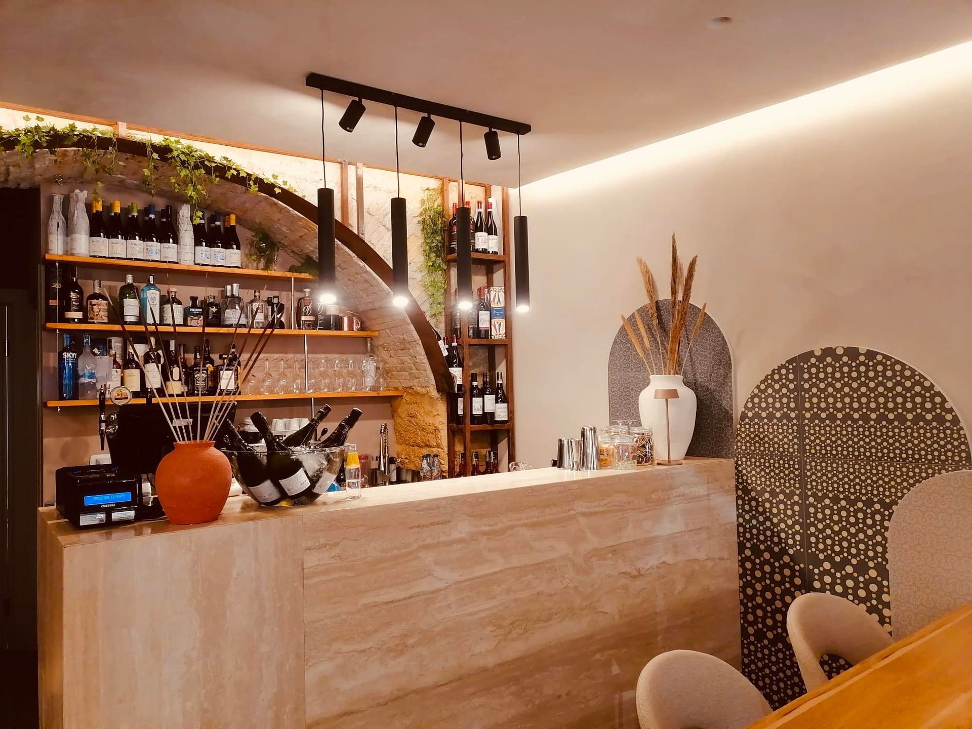 Bar con bottiglie, bevande, piante e pareti strutturate in un ambiente con luci calde.