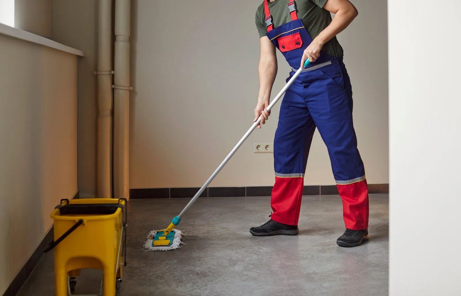 A worker in blue and red overalls mops a grey floor with a yellow bucket nearby in an indoor setting.