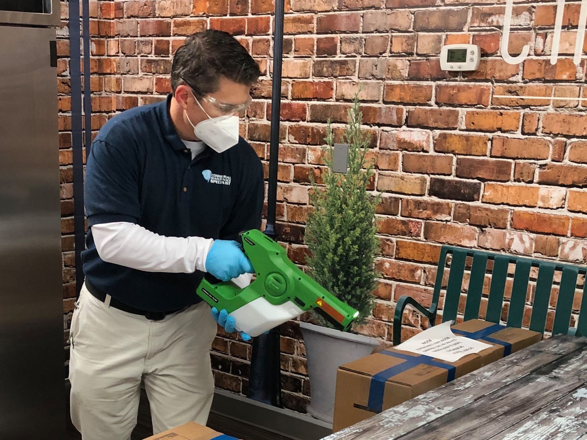 Man in PPE sanitizing with a sprayer; brick wall background, boxes, green bench.
