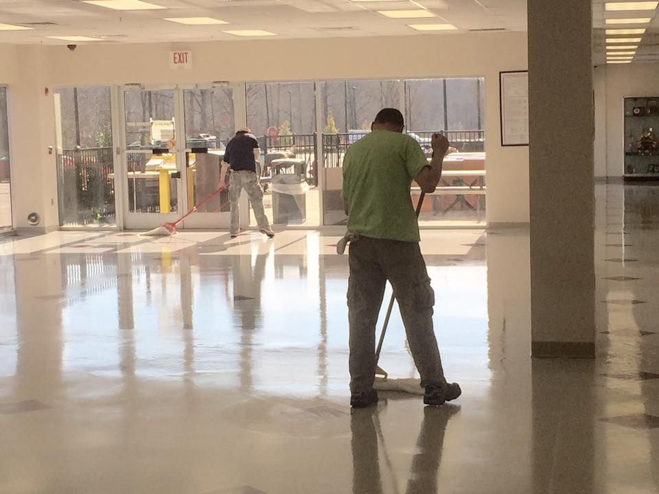 Two people mopping a large, shiny floor in a building with glass doors and bright lighting.