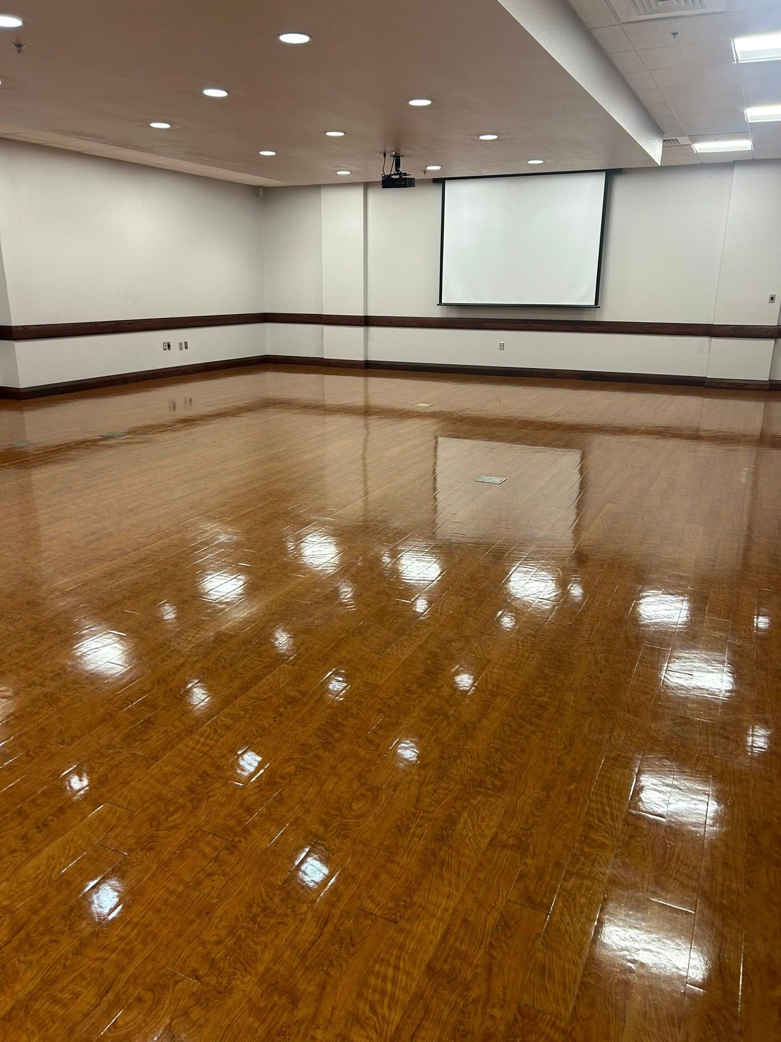 Shiny wooden floor in a large empty room with a projector screen and lights on the ceiling.