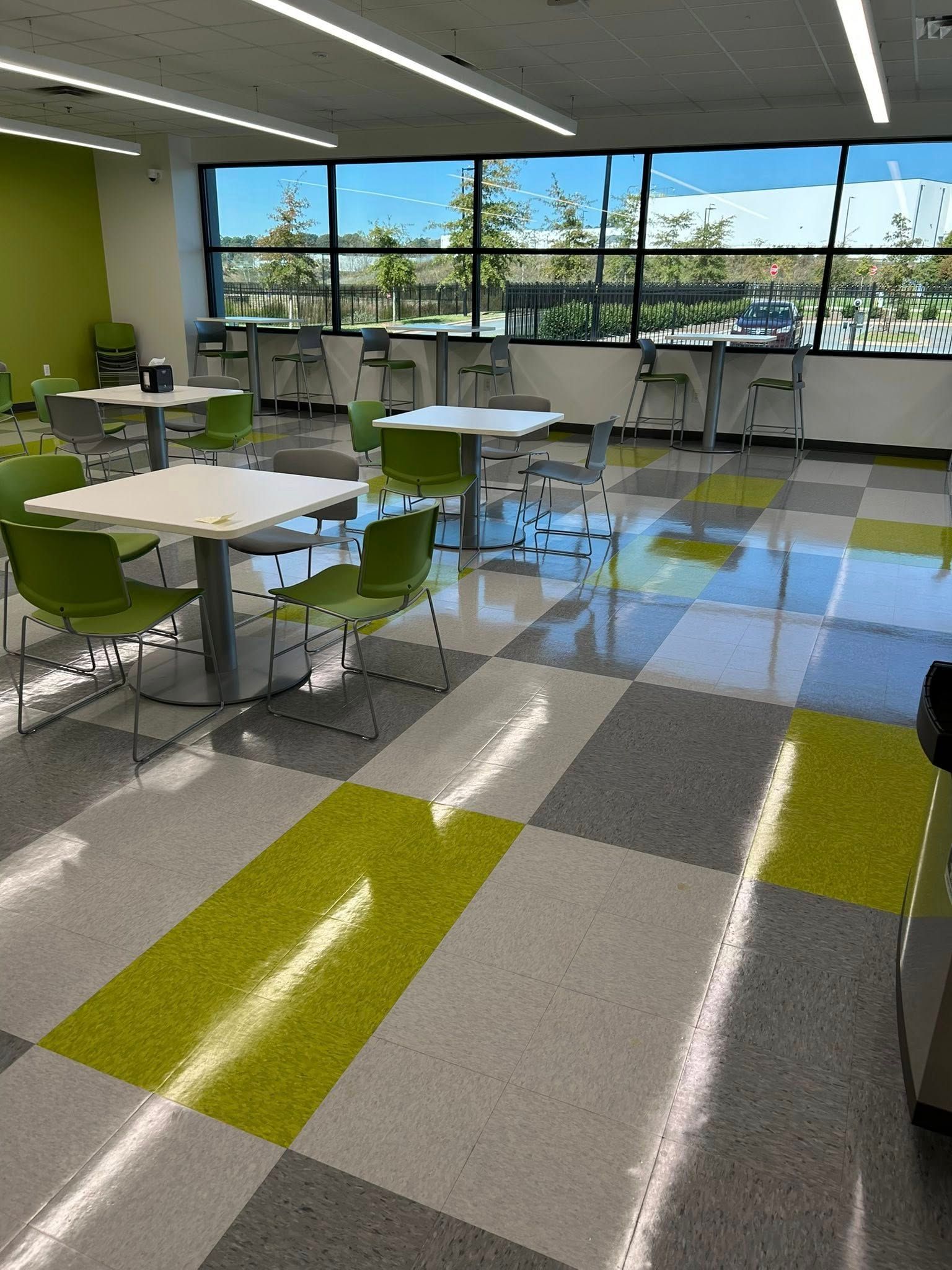 Bright cafeteria with tables, green chairs, checkered floor, large windows.