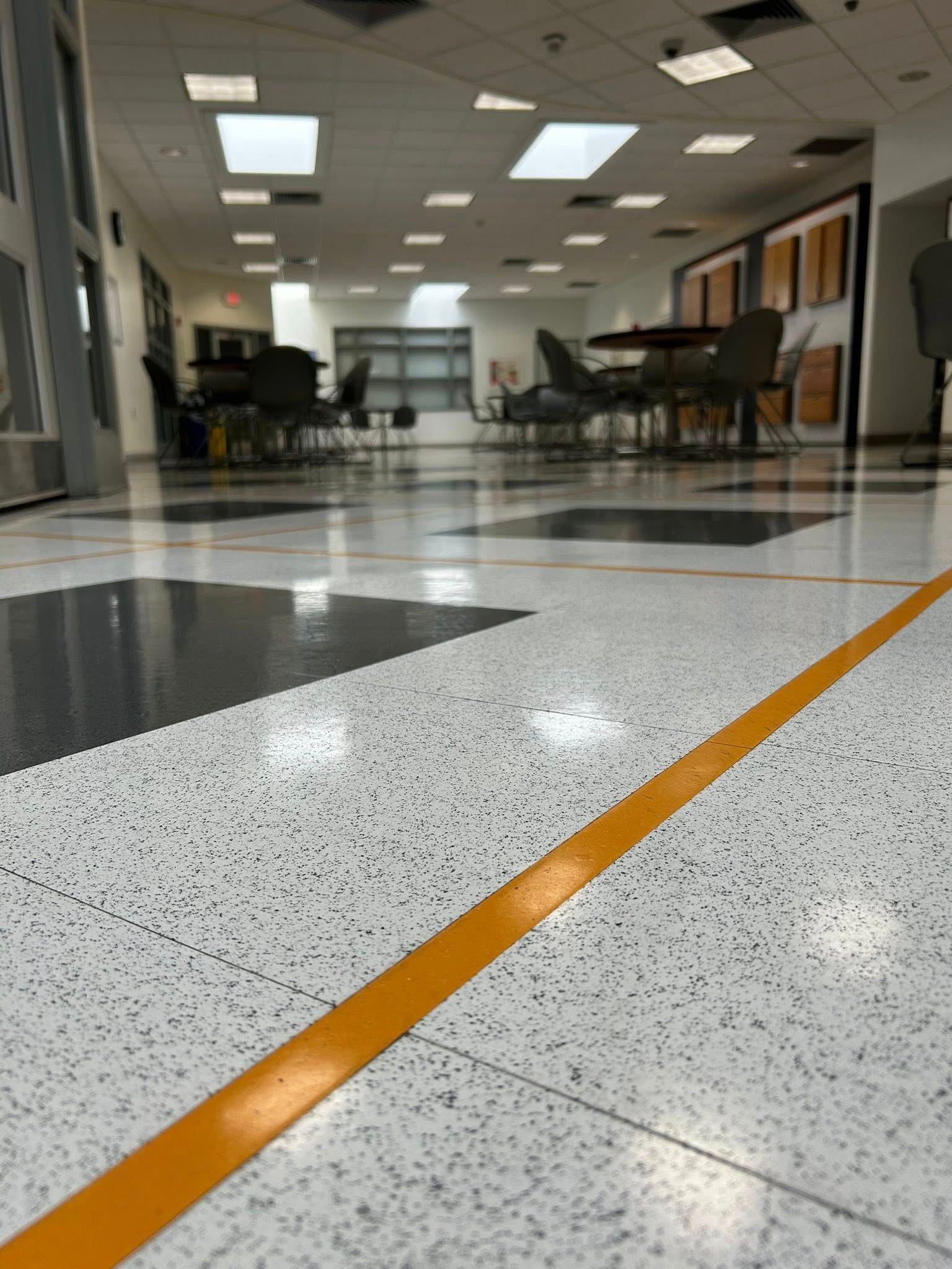 A low-angle view of a shiny floor with an orange stripe, leading to a cafeteria with tables and chairs.