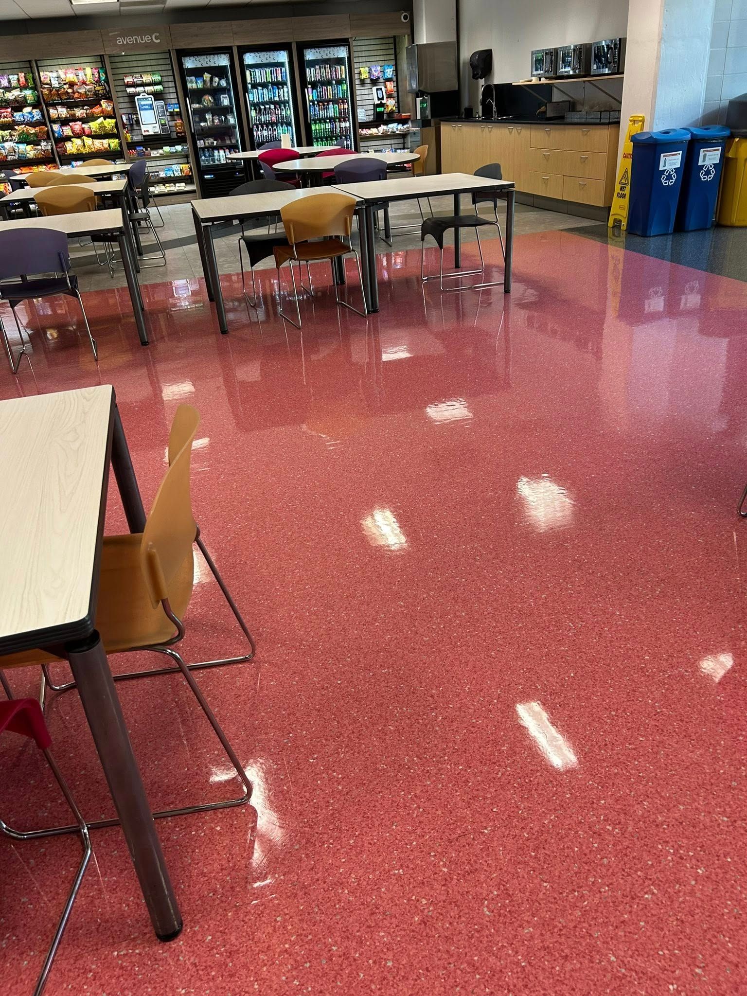 A cafeteria with red speckled floor, tables, chairs, vending machines, and trash/recycling bins.