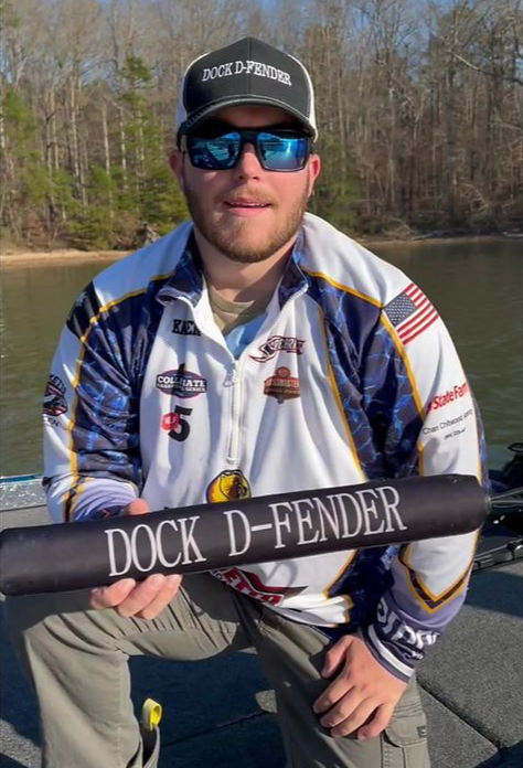 Man holding a Dock D-Fender banner, on a boat. He wears sunglasses and a fishing shirt. Water and trees in the background.
