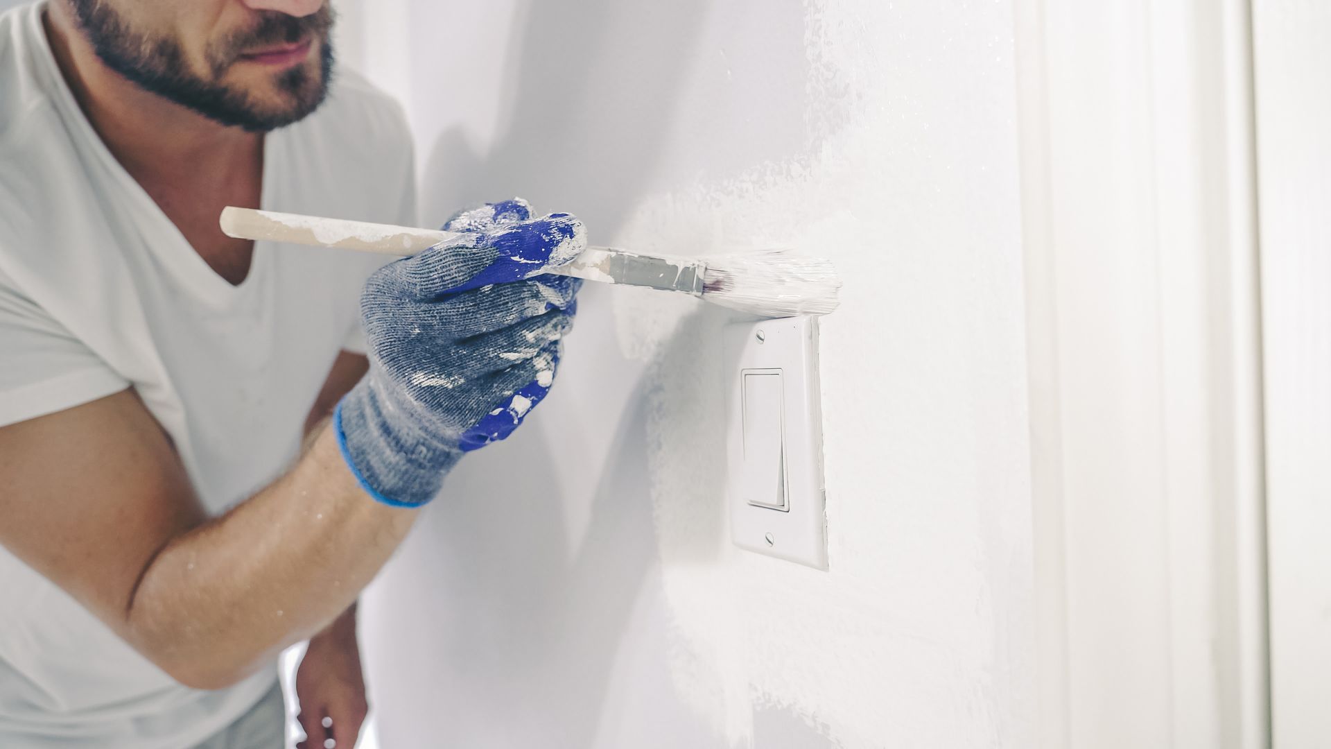 A person wearing blue gloves uses a brush to paint a white wall around a light switch plate.